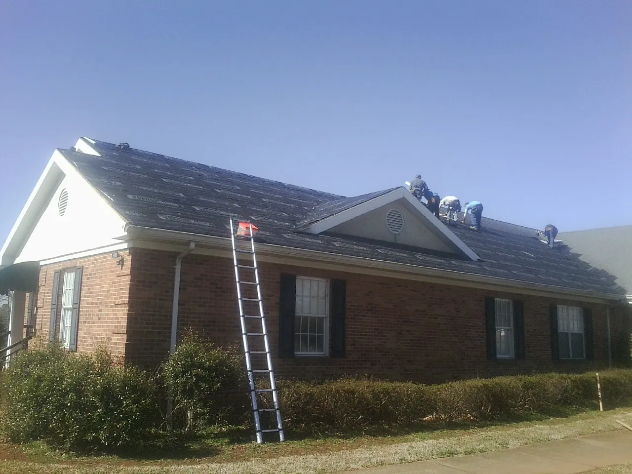 Crew tearing shingles off a brick ranch roof with a ladder set — Emergency Roof Repair work by our crew in Interlachen